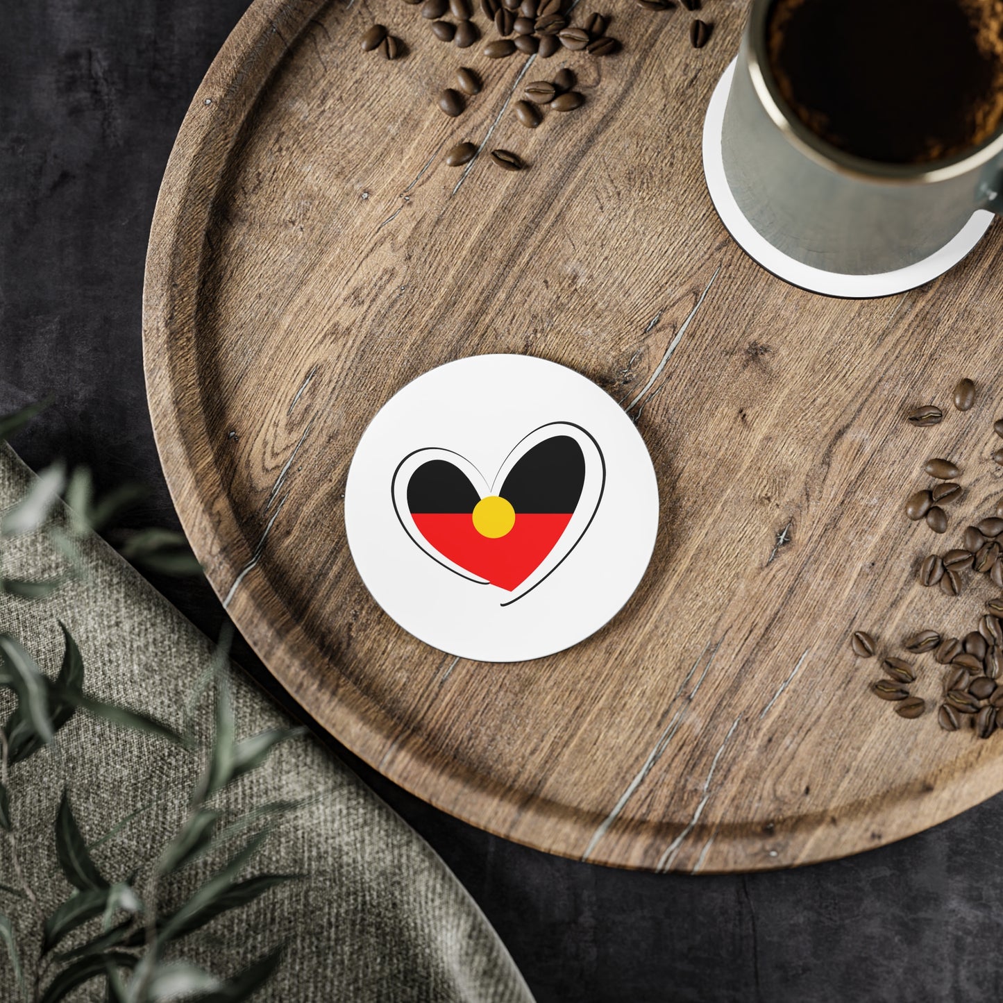 Round white Aboriginal Flag coaster placed on wooden serving board beside a coffee cup.