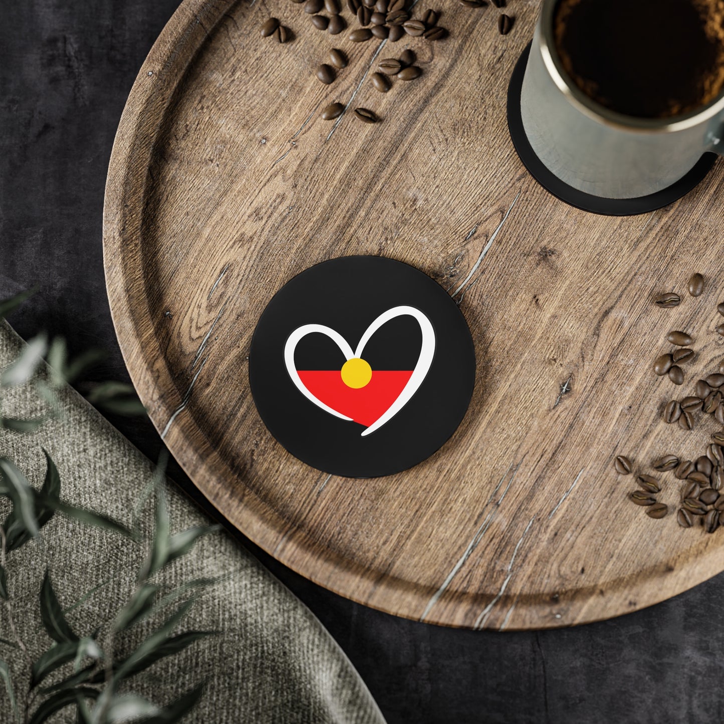Round black Aboriginal Flag coaster placed on wooden serving board beside a coffee cup.